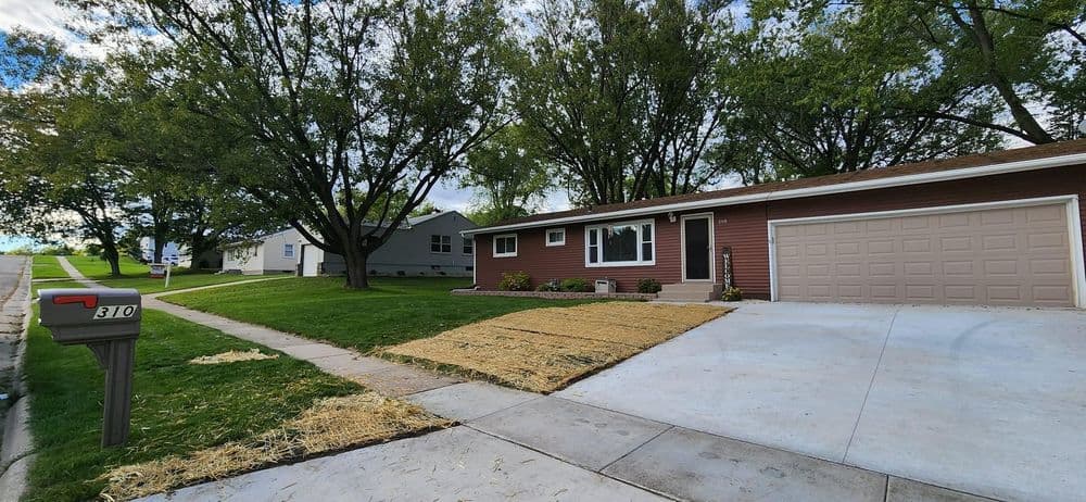 Single-story red house with garage, landscaped front yard, and tree-lined street.