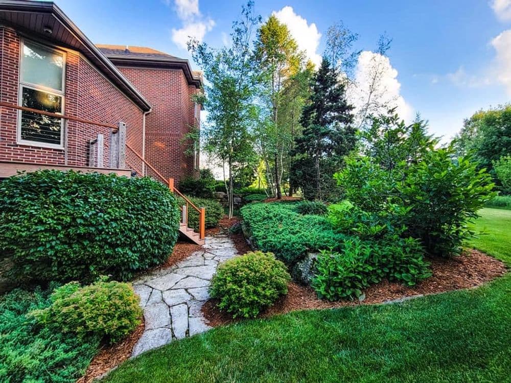 Lush garden with stone pathway leading to a brick house under a clear blue sky.
