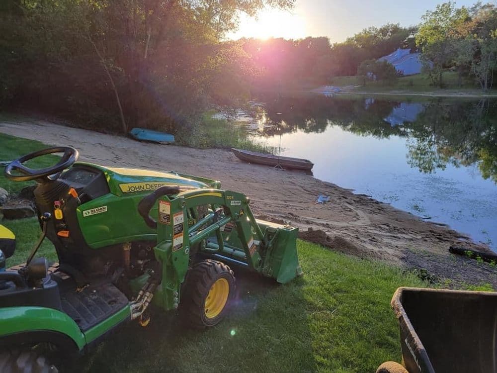 John Deere tractor by peaceful lakeside at sunset with boats in the background.