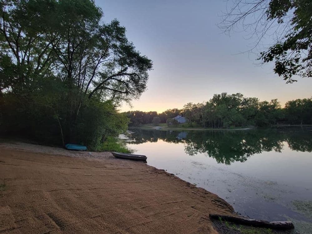 Serene lakeside view at sunrise with trees and a sandy shore reflecting calm water.