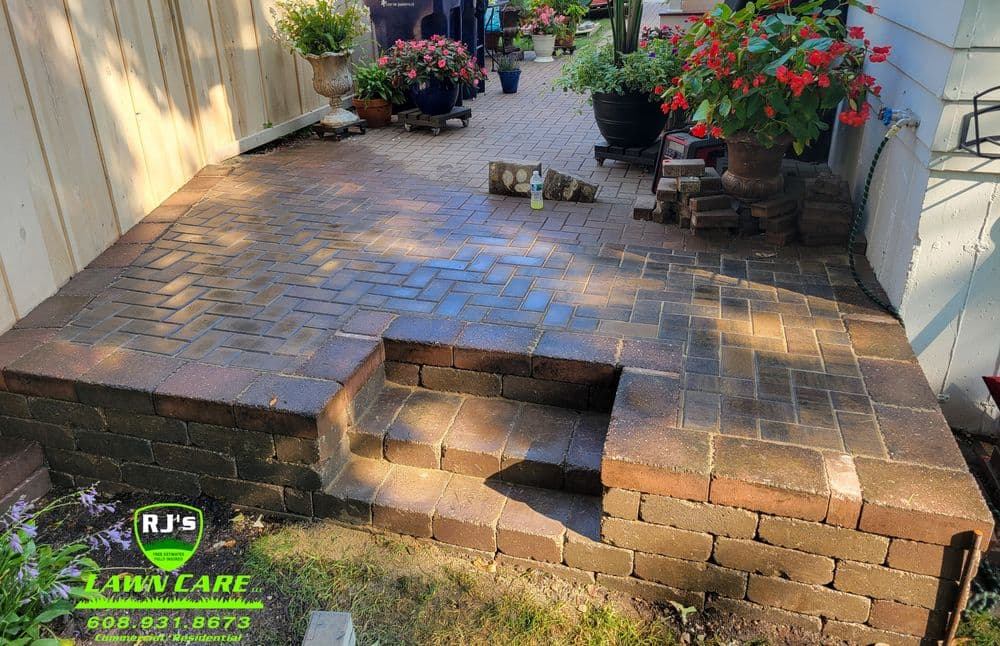 Paved patio with steps, surrounded by vibrant flower pots and greenery. RJ's Lawn Care logo.