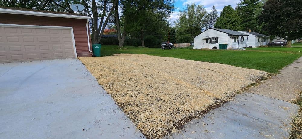 Newly prepared lawn area with straw on a residential property, adjacent to a garage and house.