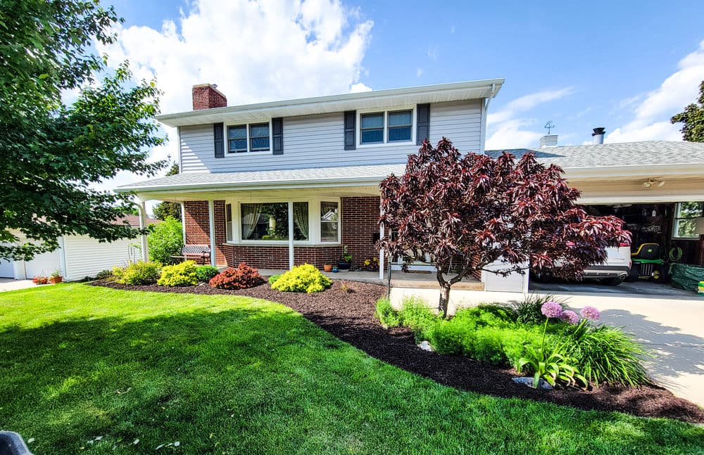Front yard of a two-story house with landscaped garden, colorful flowers, and a decorative tree.