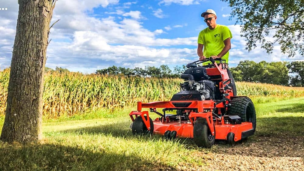 Man using a commercial lawn mower on a grassy path beside cornfields under a blue sky.