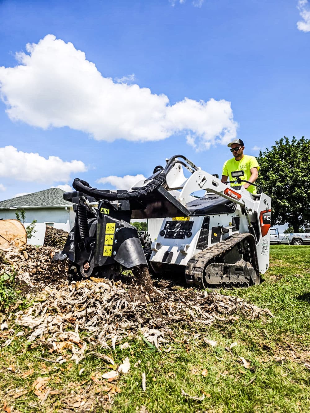 Man operating a stump grinder machine in a grassy area under a blue sky with fluffy clouds.