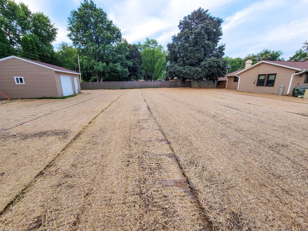 Bare soil field with straw covering, surrounded by houses and trees under a clear sky.