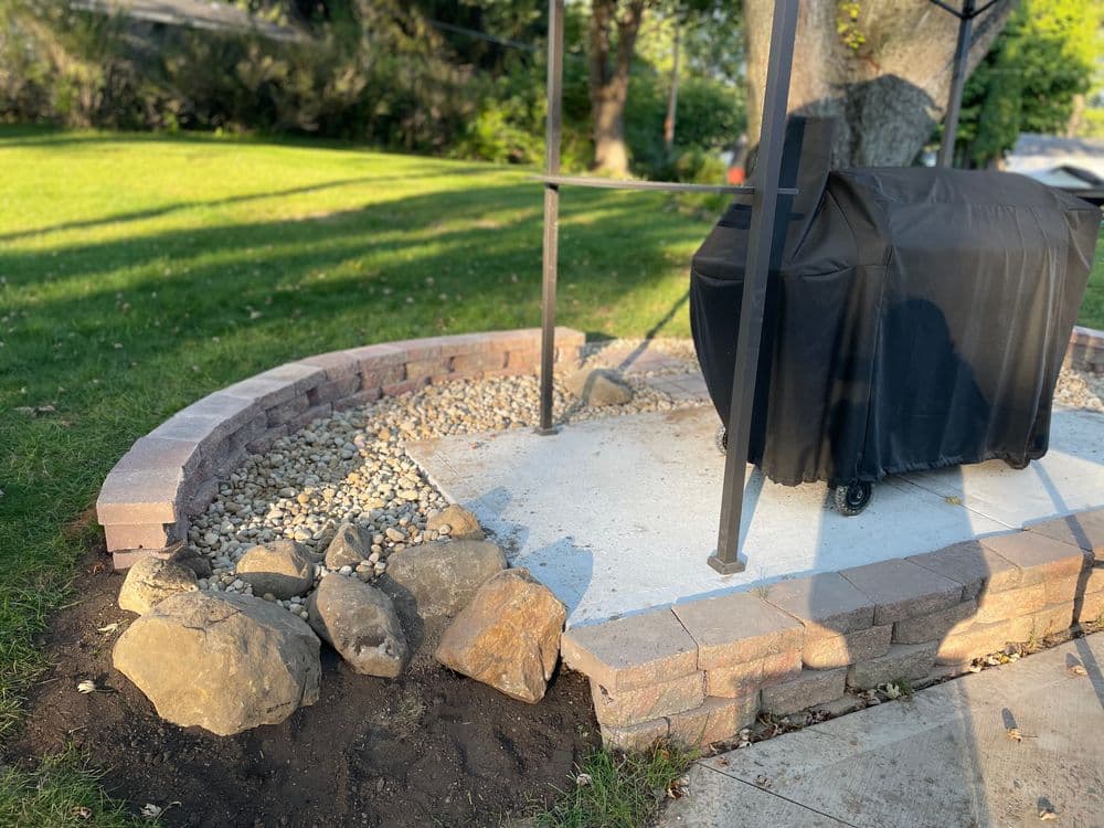 Grill covered with black tarp on a stone patio surrounded by gravel and landscaping rocks.