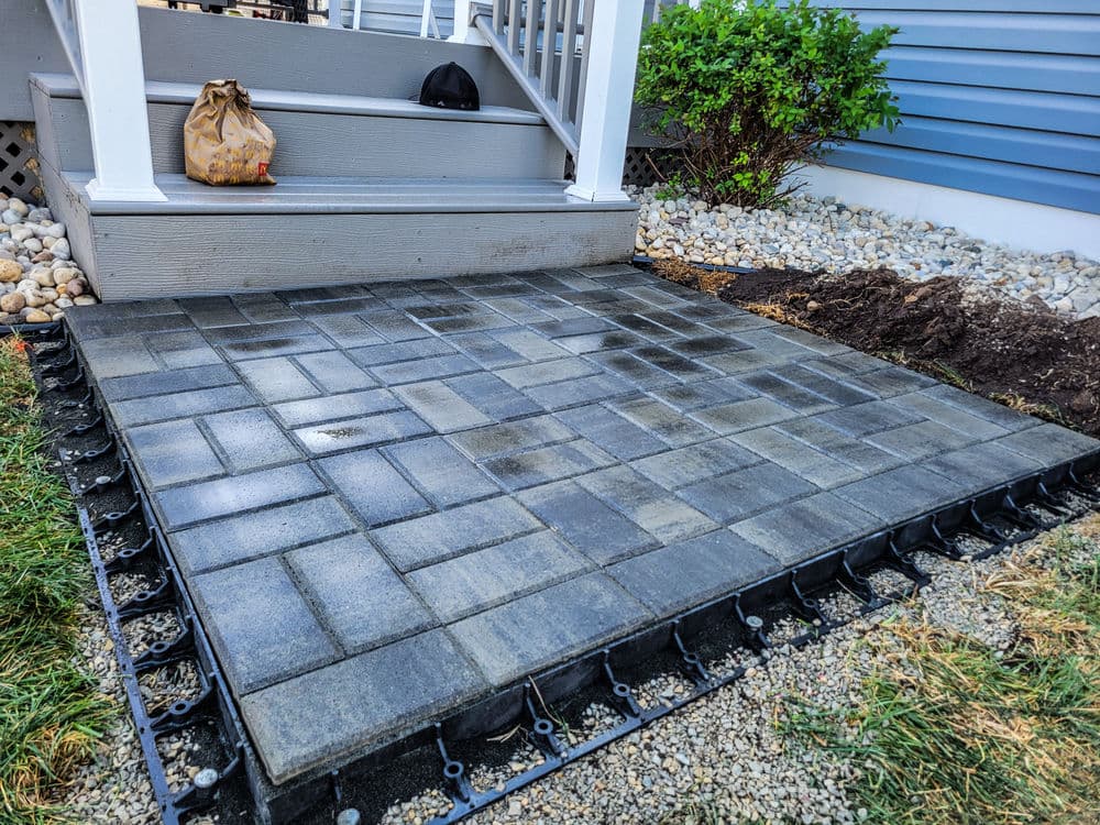 Newly installed paver patio with gray stone blocks beside a landscaped area and porch steps.