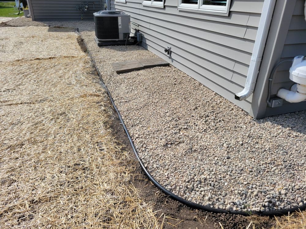 Gravel landscaping beside a house with air conditioning unit and drainage system installed.