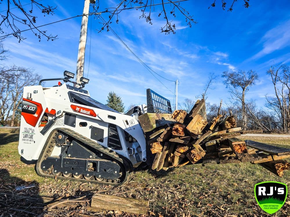 Bobcat MT100 track loader beside a pile of firewood on a sunny day. RJ’s logo visible.