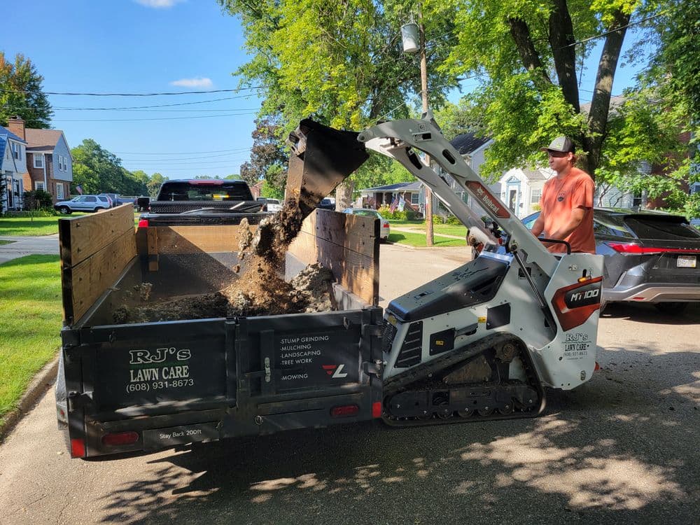 Lawn care worker using a mini skid steer to unload dirt into a truck on a residential street.