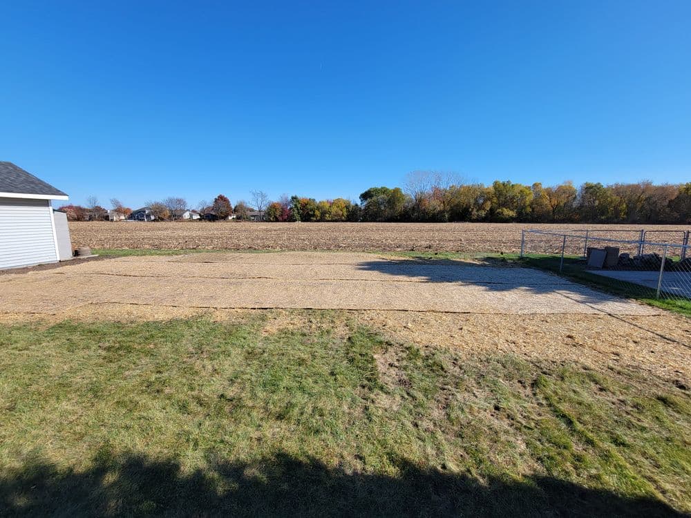 Cleared land with grass and gravel under a clear blue sky, surrounded by autumn trees.